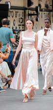Two models walking on a fashion show runway wearing light-colored outfits.