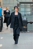 Model wearing a black outfit walking on a runway with a blurred industrial background.