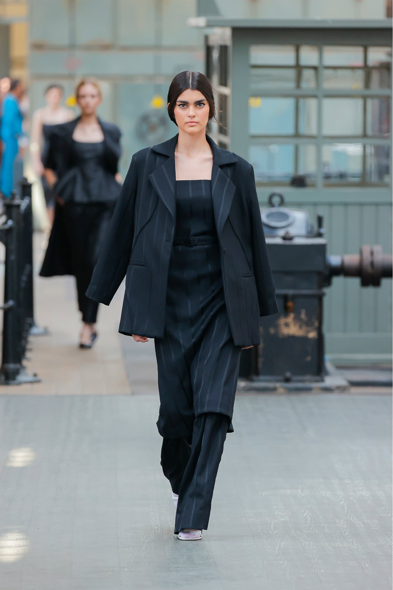 Model wearing a black outfit walking on a runway with a blurred industrial background.