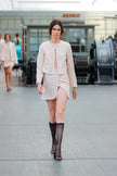 Model walking on a runway wearing a beige outfit with a blurred indoor background