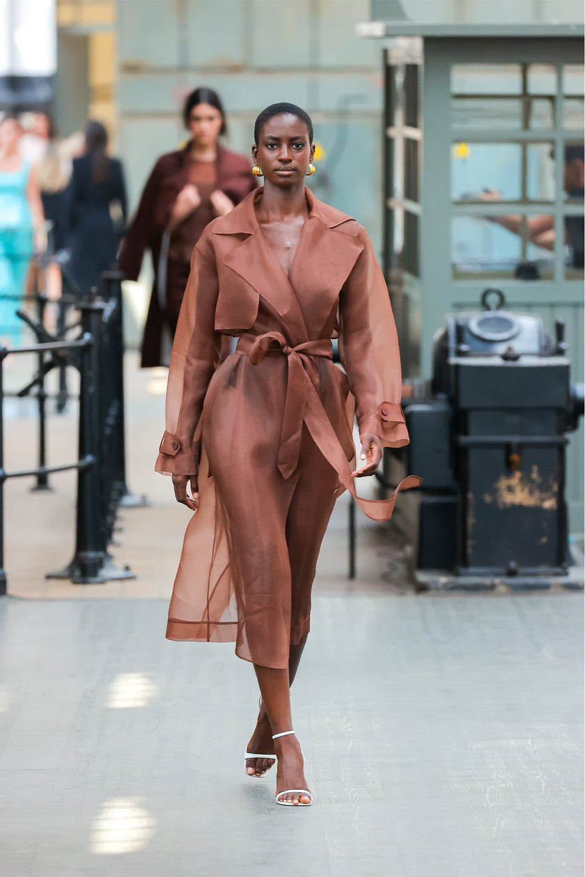 Woman in a brown trench coat walking in an indoor setting with blurred background